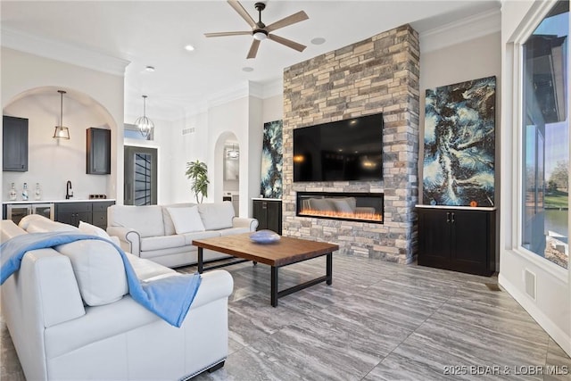 living room with a stone fireplace, ceiling fan, ornamental molding, and sink