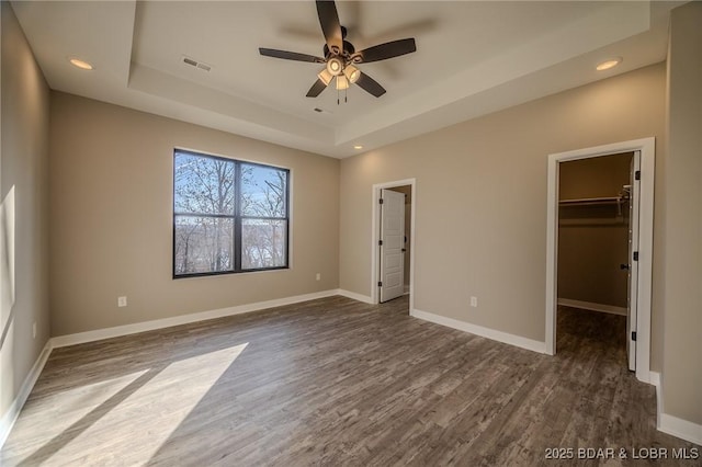 unfurnished bedroom with dark wood-type flooring, a walk in closet, ceiling fan, a tray ceiling, and a closet