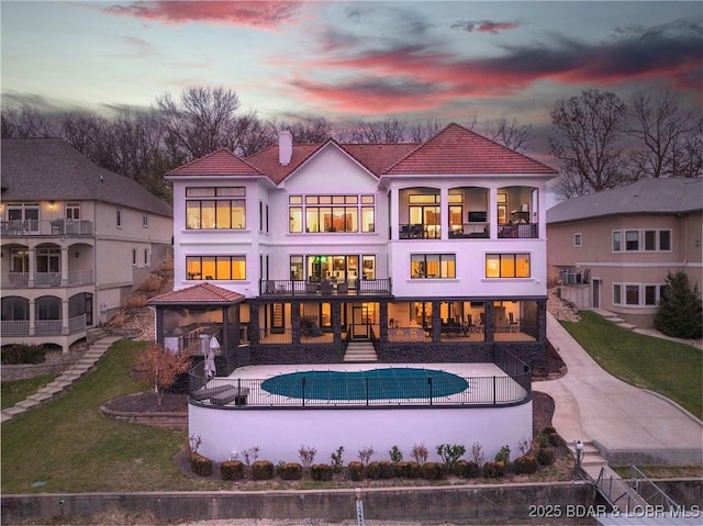 back of house at dusk featuring a chimney, stucco siding, a patio, a balcony, and a lawn