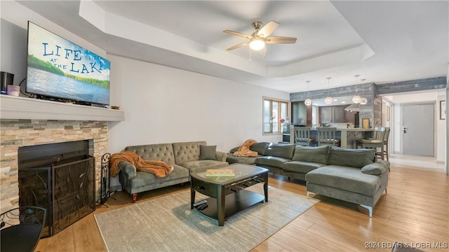 living room featuring a tray ceiling, a stone fireplace, ceiling fan, and light hardwood / wood-style floors