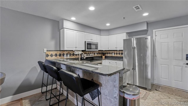 kitchen with kitchen peninsula, decorative backsplash, white cabinetry, appliances with stainless steel finishes, and dark stone counters