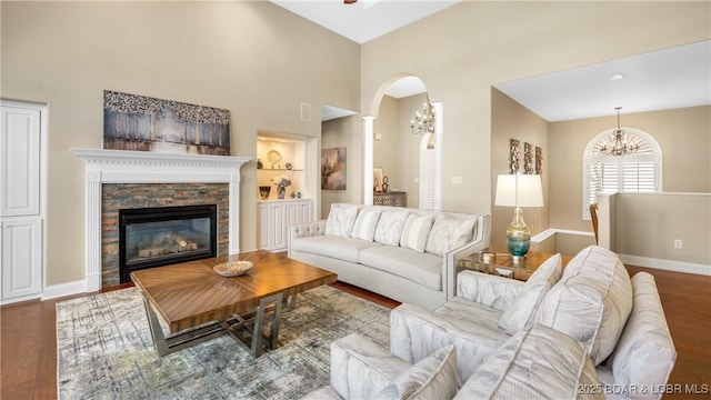 living room with built in shelves, a stone fireplace, wood-type flooring, a notable chandelier, and a towering ceiling