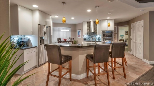 kitchen featuring wall chimney range hood, hanging light fixtures, appliances with stainless steel finishes, tasteful backsplash, and white cabinetry