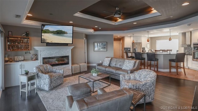 living room with ornamental molding, a raised ceiling, ceiling fan, hardwood / wood-style flooring, and a stone fireplace