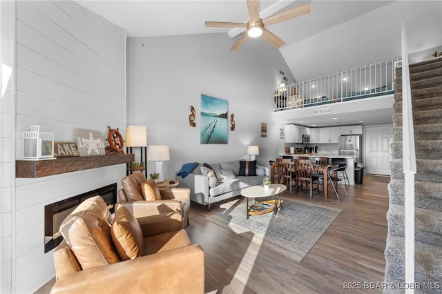 living room featuring stairway, a glass covered fireplace, ceiling fan, wood finished floors, and high vaulted ceiling