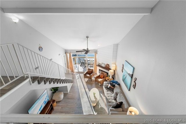 living area with lofted ceiling with beams, wood finished floors, a ceiling fan, stairs, and a glass covered fireplace