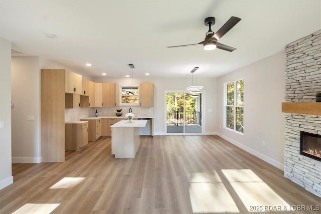 kitchen with a kitchen island with sink, ceiling fan, light brown cabinets, a stone fireplace, and hanging light fixtures