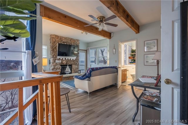 living room with beamed ceiling, a stone fireplace, ceiling fan, and light hardwood / wood-style flooring