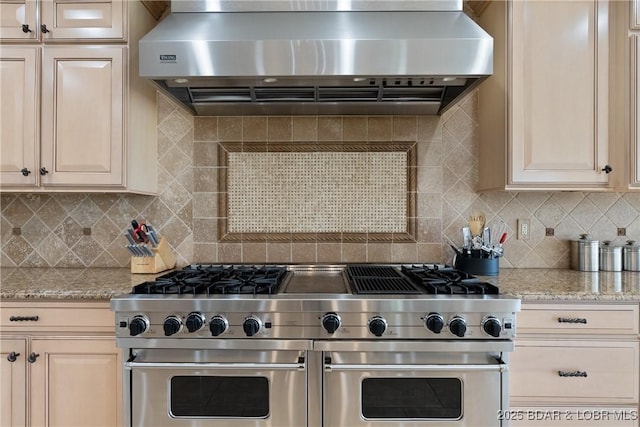 kitchen with light stone counters, double oven range, wall chimney range hood, and backsplash