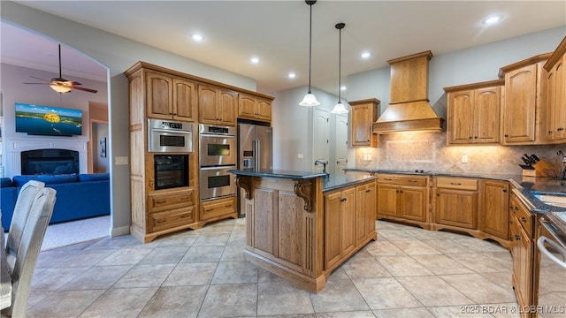 kitchen featuring appliances with stainless steel finishes, backsplash, custom exhaust hood, hanging light fixtures, and an island with sink