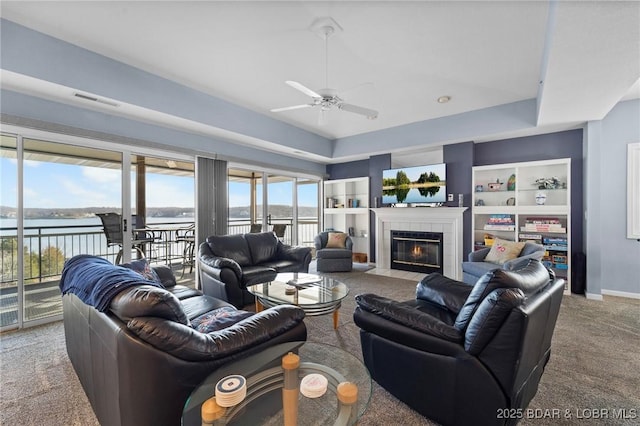 carpeted living room featuring a tile fireplace, a water view, ceiling fan, a tray ceiling, and built in shelves