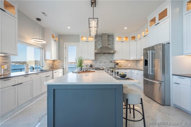 kitchen featuring wall chimney range hood, an island with sink, hanging light fixtures, a water view, and high end fridge