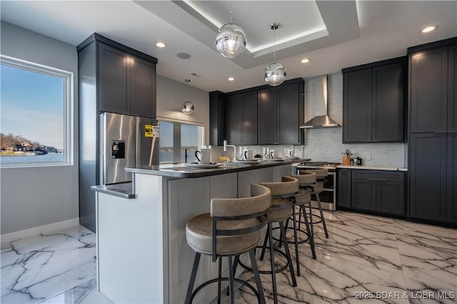 kitchen with wall chimney exhaust hood, decorative light fixtures, a kitchen bar, stainless steel appliances, and a tray ceiling