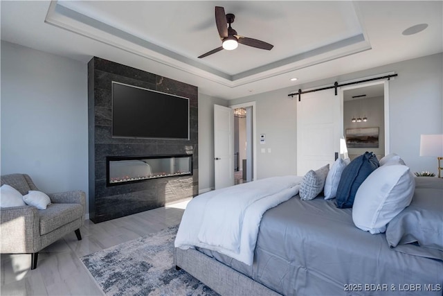 bedroom with ceiling fan, a barn door, light hardwood / wood-style floors, a tray ceiling, and a tiled fireplace