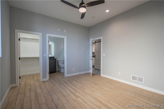 unfurnished bedroom with ceiling fan, ensuite bathroom, a barn door, and light wood-type flooring