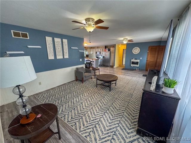 living area featuring heating unit, visible vents, a ceiling fan, wainscoting, and wood finished floors