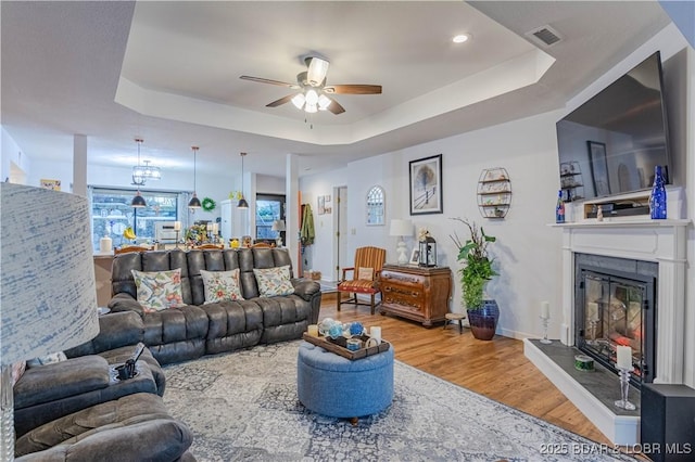 living area featuring wood finished floors, a tray ceiling, visible vents, ceiling fan, and a glass covered fireplace