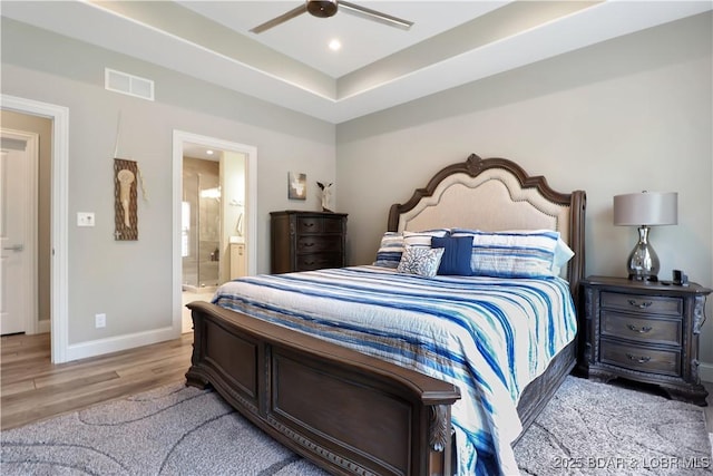 bedroom with light wood-type flooring, connected bathroom, a tray ceiling, and ceiling fan