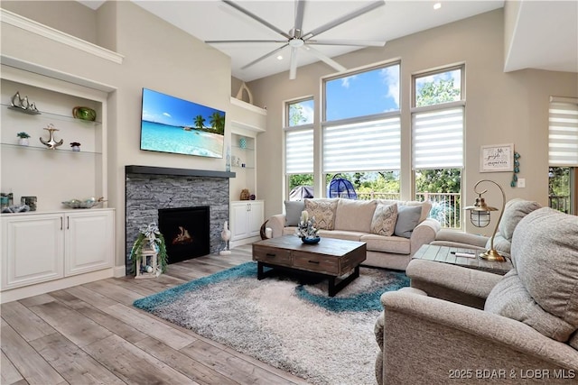 living room with ceiling fan, built in features, a stone fireplace, and light wood-type flooring