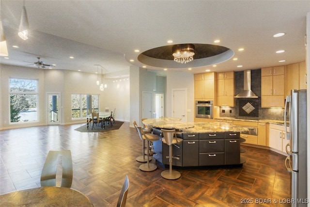 kitchen with a center island, appliances with stainless steel finishes, light stone counters, wall chimney exhaust hood, and light brown cabinets