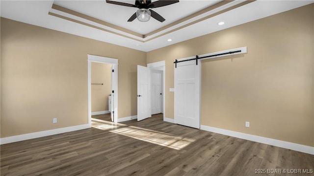 unfurnished bedroom with dark wood-type flooring, a tray ceiling, ceiling fan, and a barn door