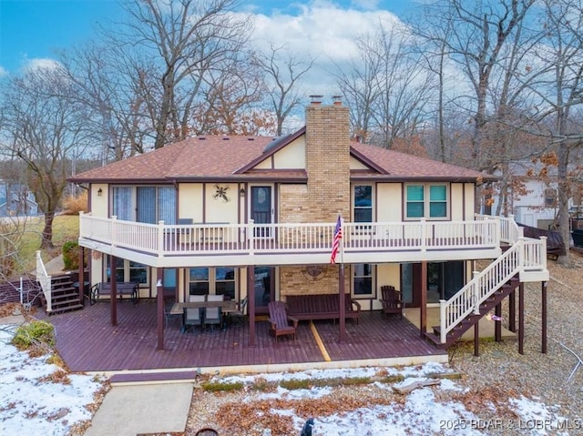 snow covered back of property featuring a chimney, roof with shingles, stairs, a deck, and stucco siding