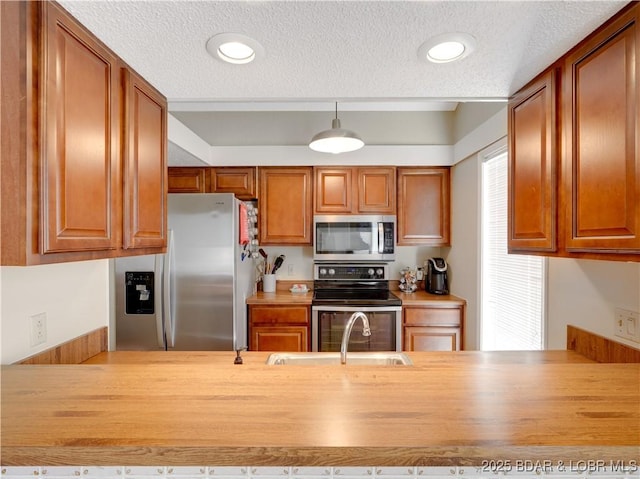kitchen with brown cabinetry, a textured ceiling, a sink, appliances with stainless steel finishes, and decorative light fixtures