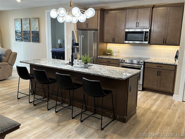 kitchen with an island with sink, appliances with stainless steel finishes, light stone counters, and light hardwood / wood-style floors