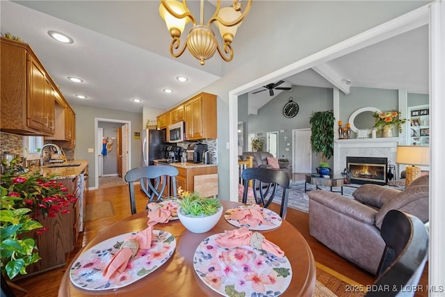 dining area featuring ceiling fan with notable chandelier, wood-type flooring, vaulted ceiling with beams, sink, and a premium fireplace