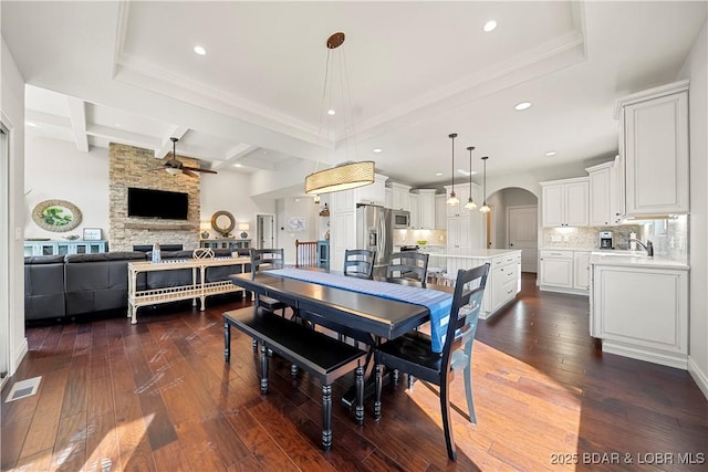 dining area with dark hardwood / wood-style flooring, a stone fireplace, beamed ceiling, and ceiling fan