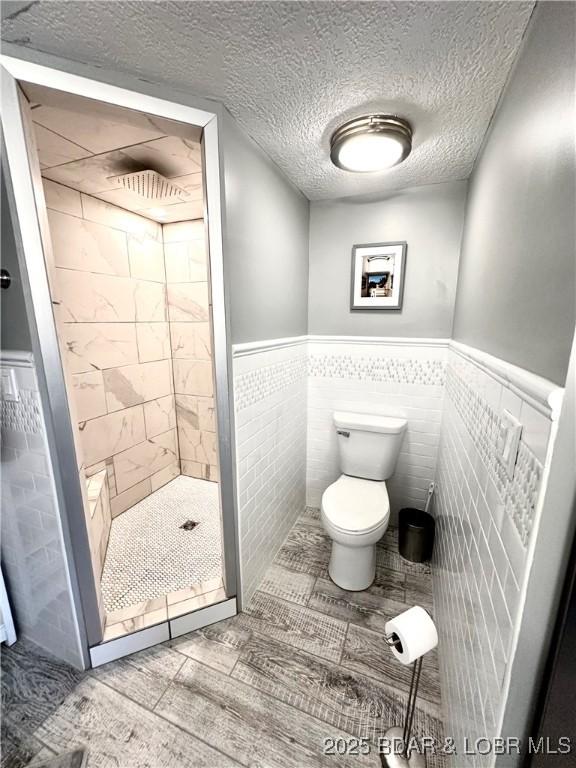 bathroom with toilet, wood-type flooring, a textured ceiling, tile walls, and a tile shower