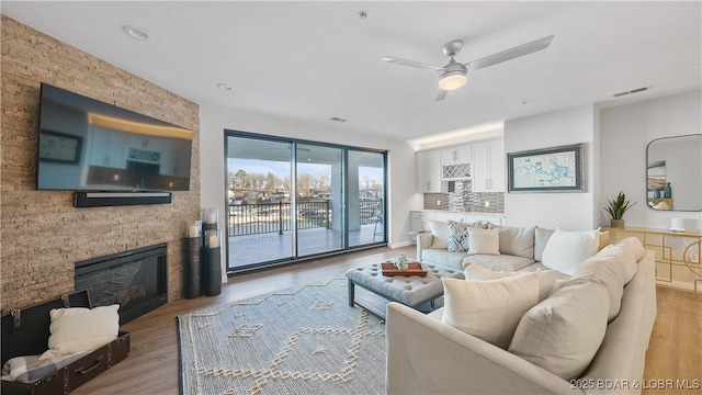 living area featuring a stone fireplace, light wood-type flooring, visible vents, and a ceiling fan