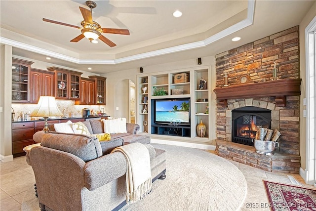 tiled living room featuring a stone fireplace, a raised ceiling, and ceiling fan