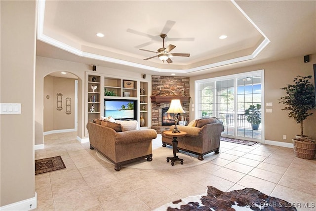 tiled living room featuring ceiling fan, a tray ceiling, and a stone fireplace