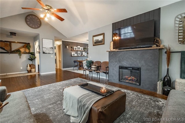 living area featuring baseboards, a glass covered fireplace, ceiling fan, wood finished floors, and high vaulted ceiling