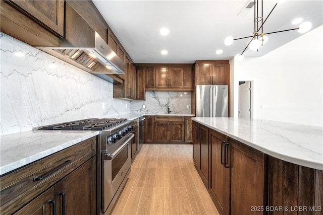 kitchen with tasteful backsplash, light wood-type flooring, premium appliances, light stone countertops, and wall chimney range hood