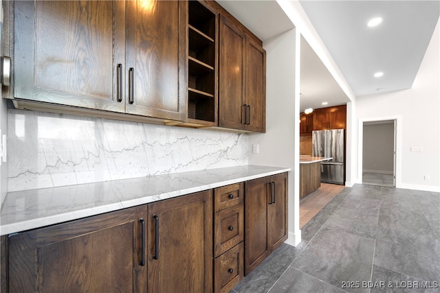 kitchen featuring light stone counters, dark brown cabinetry, stainless steel fridge, and decorative backsplash