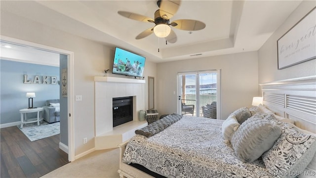 bedroom with a fireplace, wood-type flooring, access to outside, ceiling fan, and a raised ceiling