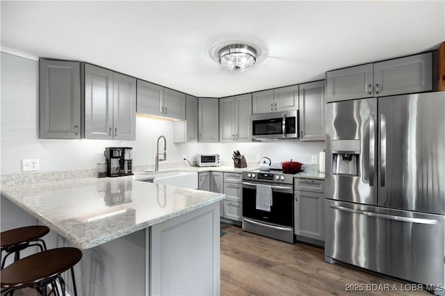 kitchen with gray cabinetry, appliances with stainless steel finishes, a sink, light stone countertops, and a peninsula