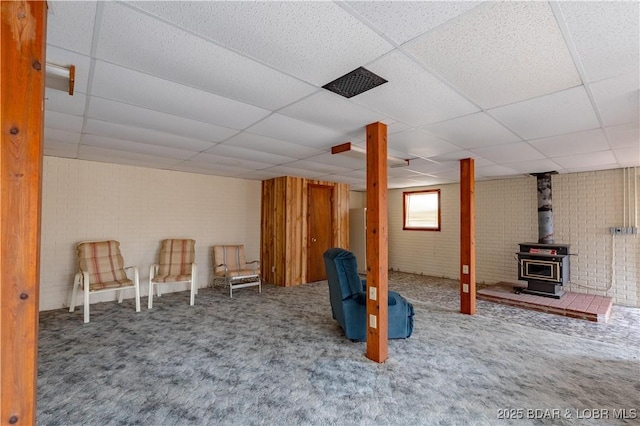 basement with a wood stove, carpet floors, a paneled ceiling, and brick wall