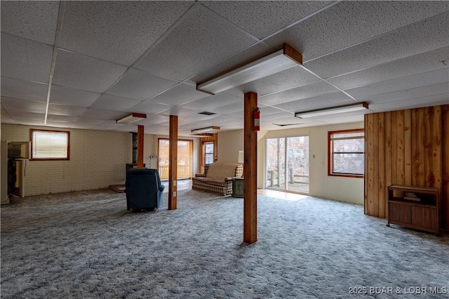 basement featuring brick wall, carpet, a wood stove, and a paneled ceiling