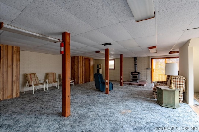 basement featuring carpet, a wood stove, a paneled ceiling, and plenty of natural light