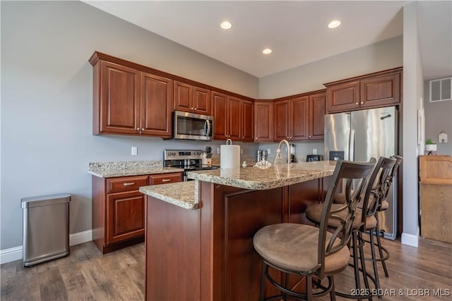 kitchen with light stone counters, stainless steel appliances, dark hardwood / wood-style floors, and a center island with sink