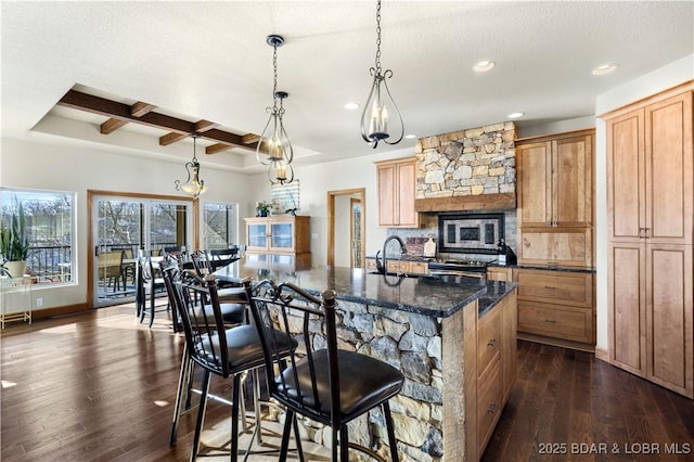 kitchen with an island with sink, coffered ceiling, dark wood finished floors, and decorative light fixtures