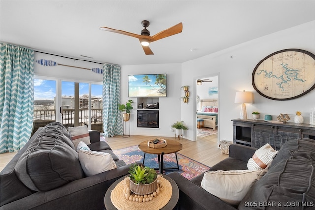 living area with ceiling fan, a glass covered fireplace, and light wood-style flooring