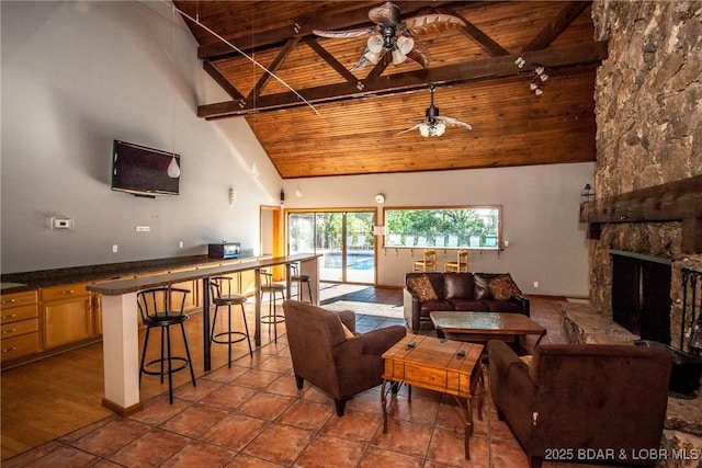 tiled living room featuring a ceiling fan, wood ceiling, beamed ceiling, a stone fireplace, and high vaulted ceiling