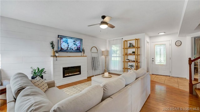 living area with stairs, ceiling fan, a glass covered fireplace, and light wood-style floors