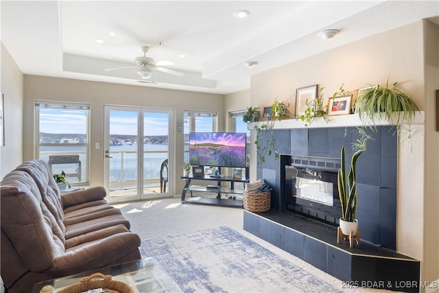 living area with carpet floors, a tray ceiling, a tiled fireplace, and a ceiling fan