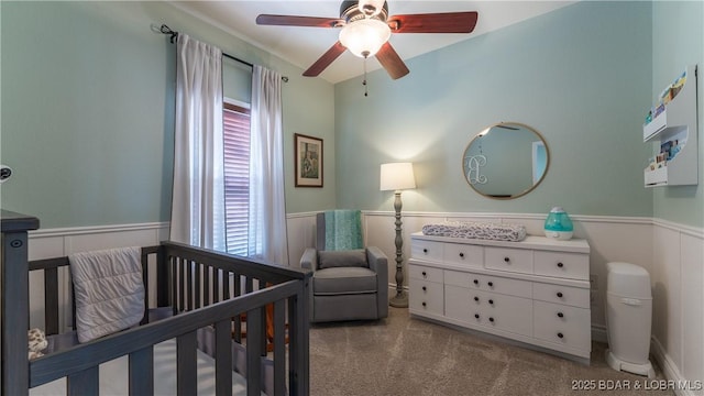 carpeted bedroom featuring a wainscoted wall, ceiling fan, and a crib