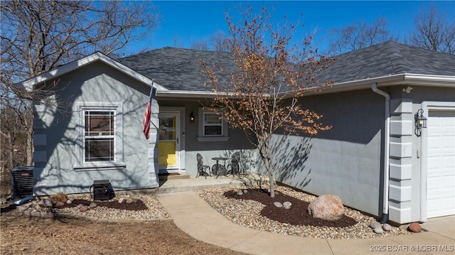 ranch-style house featuring an attached garage, central air condition unit, a shingled roof, and stucco siding